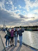 Die vier Schülerinnen stehen vor einem Hafen. Im Hintergrund sind Boote und kleine Schiffe zu sehen.