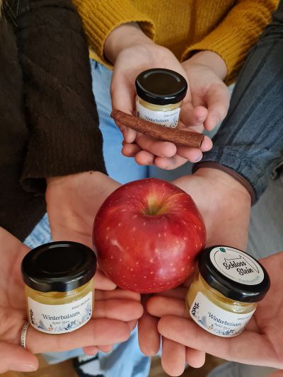 Drei Personen halten den Winterbalsam, eine Zimtstange und einen Apfel in den Händen.