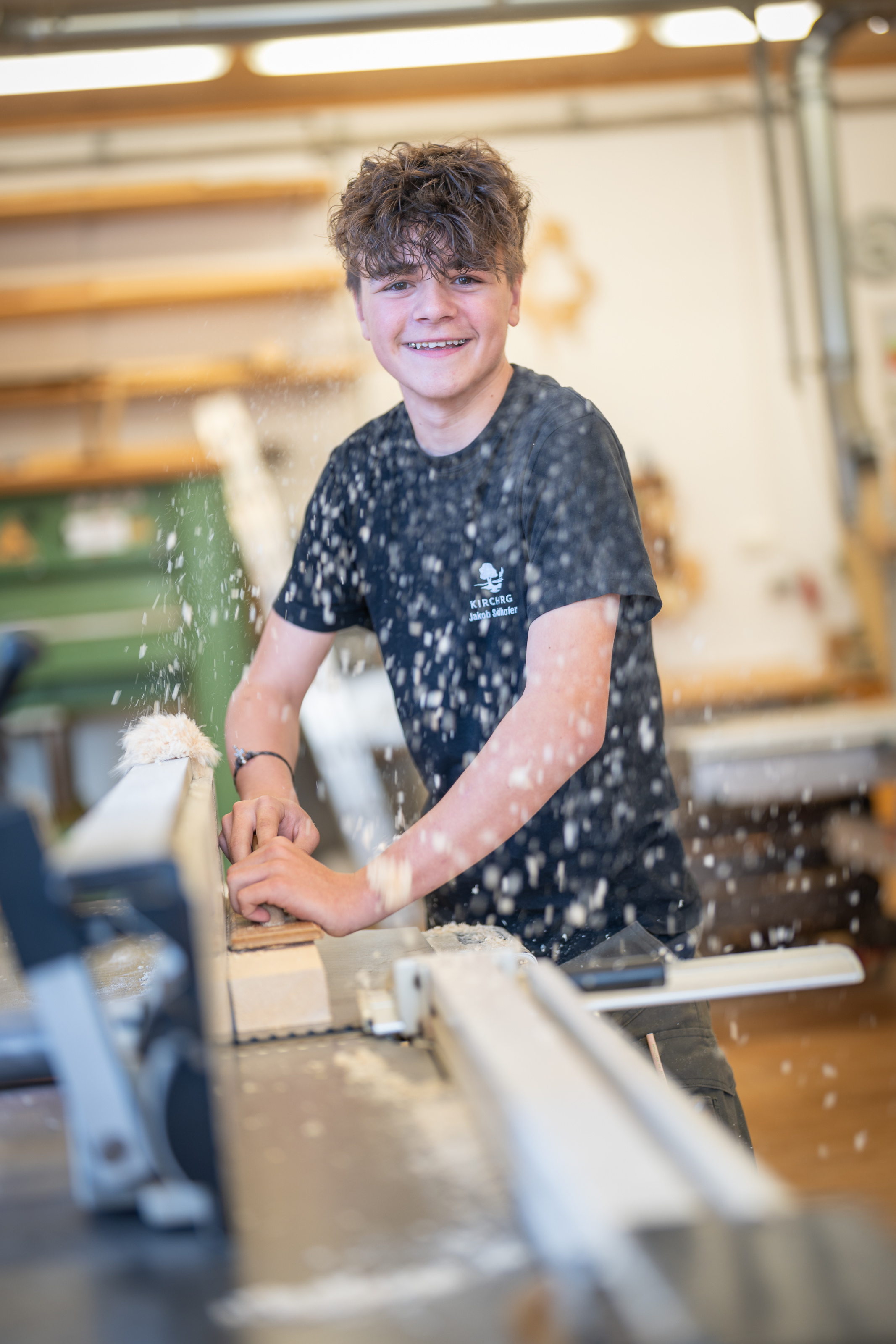 Am Foto sieht man einen männlichen Schüler, der an der Hobelmaschine steht und ein Holzstück hobelt. Er blickt freundlich lächelnd in die Kamera