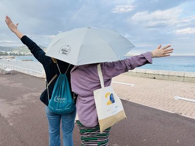 Frau Dir. Prutsch und unsere Lehrerin Frau Pregartner stehen am Meer,halten einen Regenschirm über sich und strecken die freien Hände in die Höhe. Sie tragen Taschen mit Aufdruck.