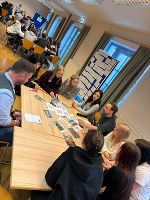 Gruppe diskutiert an einem Tisch, Fokus liegt auf ausgelegten Materialien und lebhafter Austausch.