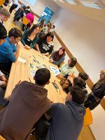 Gruppe diskutiert an einem Tisch, Fokus liegt auf ausgelegten Materialien und lebhafter Austausch.