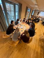 Workshop-Szene: Teilnehmende sitzen in einem Kreis, interagieren und tauschen sich aus. Helles Raumambiente.