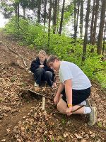 Im Wald knien sich zwei Personen über einen natürlichen Bau aus Zweigen und Laub.