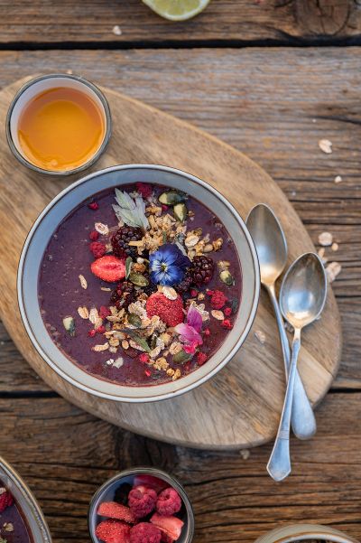 Die Aronia Bowl angerichtet in einer Schüssel, welche auf einem Holzbrett steht. Daneben liegen zwei Löffel, getrocknete Beeren und ein Getränk. Garniert ist die Bowl mit getrockneten Früchten, Granola und Blüten.