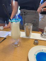 Die Schülerinnen, welche man im Hintergrund sieht, haben einen Mocktail gemixt und in einem schönen Glas, welches in Sirup und Zucker getunkt wurde, angerichtet. Daneben steht noch ein Shaker und ein Teller mit einem blauen Sirup. 