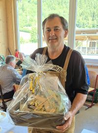 Roman Wilfing hält einen Geschenkkorb hoch. Im Hintergrund sind weitere Personen und eine helle Umgebung zu sehen.