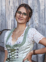 Junge Frau mit Brille und traditioneller Tracht lächelt vor einer Holzfassade.