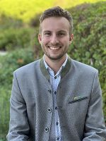 Lachende Person mit grauem Sakko vor grüner Hecke. Freundliche Ausstrahlung.