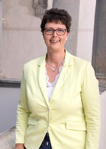 Hintergrund hellgrau, im Vordergrund junge Frau mit dunklen kurzen Haaren und einer Brille, freundlich lächelnd mit hellgelbem Blazer.
