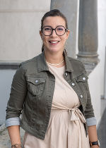 Eine Dame mit olivfarbener Jacke und hellem Kleid, dunklen zusammengemachten Haaren und einer Brille