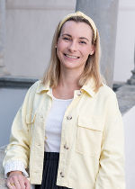 Hellgrauer Hintergrund davor eine junge Frau mit hellgelber Jacke und mittellangen blonden Haaren.