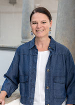 Lachende Person mit dunklem Haar, trägt Jeansjacke und weißes Oberteil. Im Hintergrund Säulen einer Gebäudeansicht. Porträtaufnahme. 
