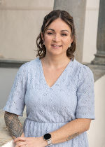 hellgrauer Hintergrund, davor eine Frau mit mittellangem dunklem Haar und hellblauem Kleid