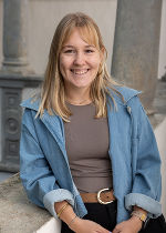 Frau mit blonden mittellangem Haar und hellblauer Jeansjacke und schlammfarbenen Shirt vor hellgrauem Hintergrund
