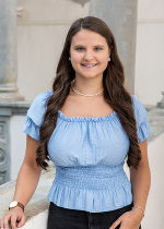 junge Frau mit langem dunklem Haar und hellblauer Bluse vor hellgrauem Hintergrund