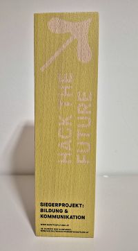 Pokal aus gelben Holzblock, dem Text „HACK THE FUTURE“ und „Siegerprojekt: Bildung & Kommunikation“.