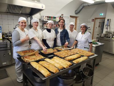 Schülerinnen, Lehrerinnen und Küchenpersonal stehen hinter einem Tisch auf dem mehrere Backbleche mit dem fertigen Gemüsewinterstrudel gemessen werden.